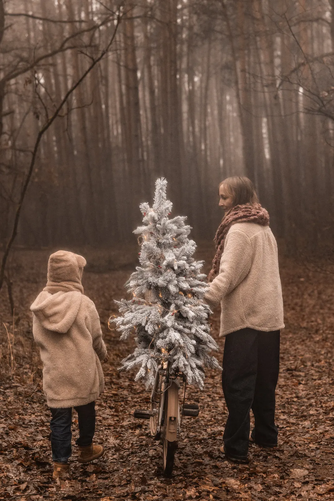 Portret matki z dzieckiem prowadzącej rower z choinką na bagażniku przez las we mgle