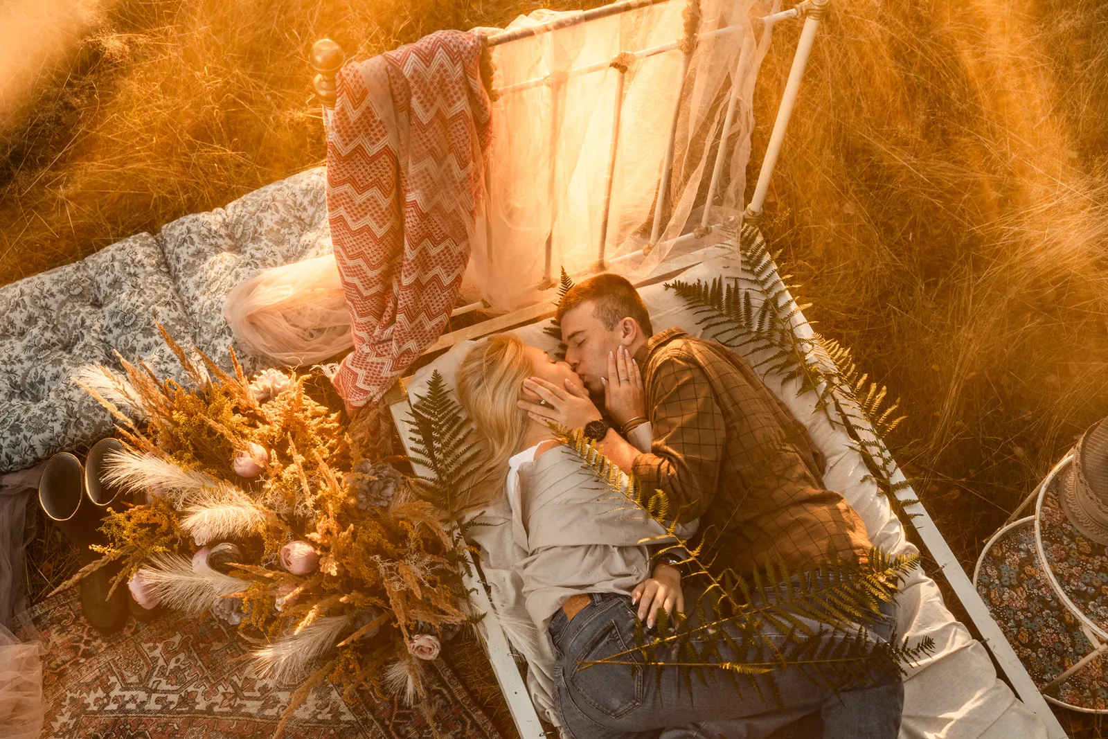 Przytulona para całuje się na łóżku w samym środku plenerowej scenerii, w tle łąka, las i oryginalna aranżacja sesji zdjęciowej