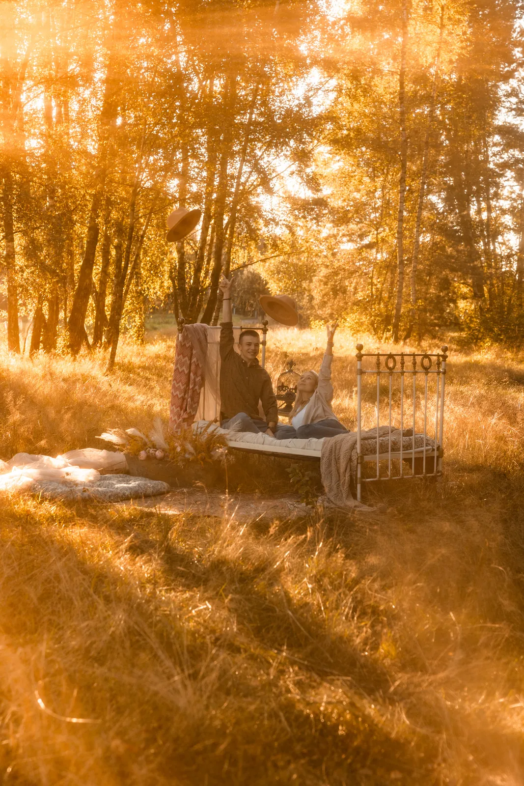 Para siedząca na łóżku retro wśród leśnej scenerii oświetlanej przez wschód słońca, podrzucają razem w górę kapelusze