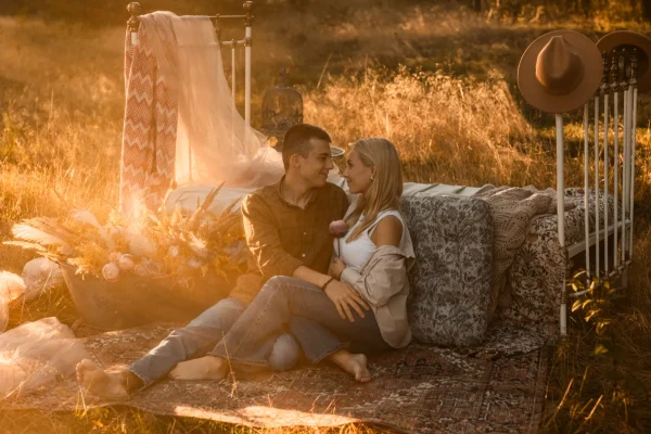 Para przytulona podczas romantycznej sesji narzeczeńskiej w plenerze o wschodzie słońca, w tle las, łóżko i kwiaty