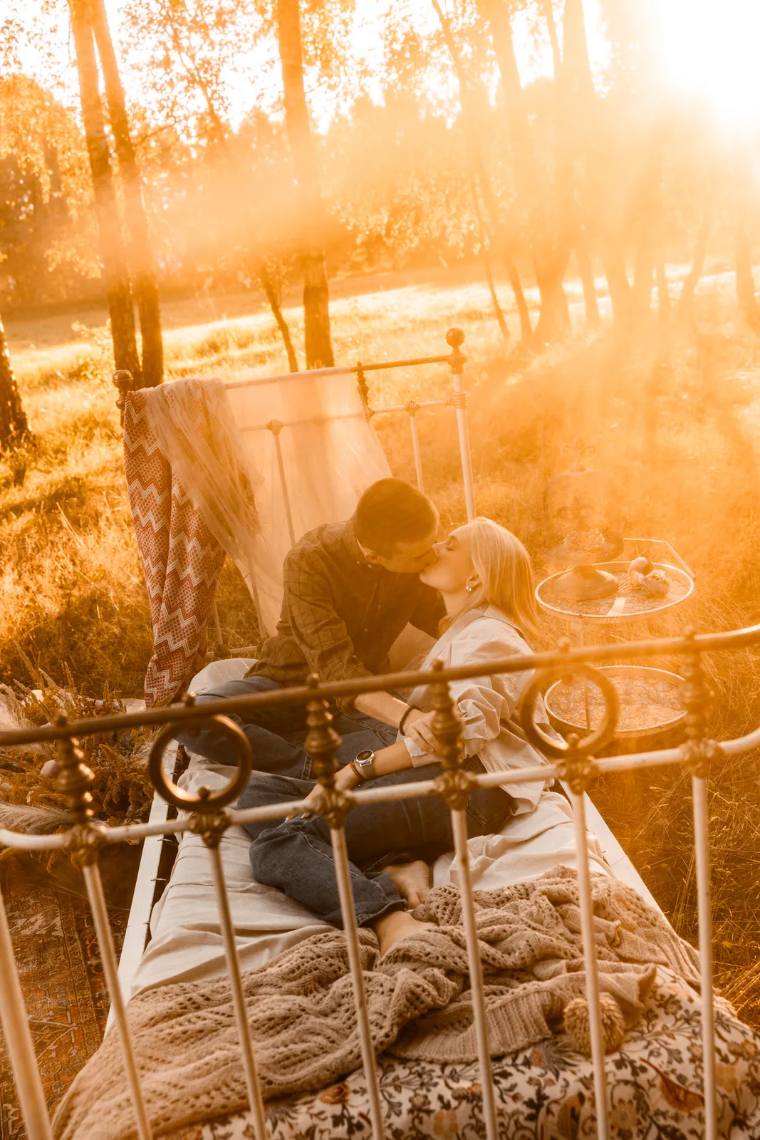 Para siedzi na łóżku i całuje się podczas romantycznej sesji narzeczeńskiej w plenerze w stylu boho w środku leśnej polany