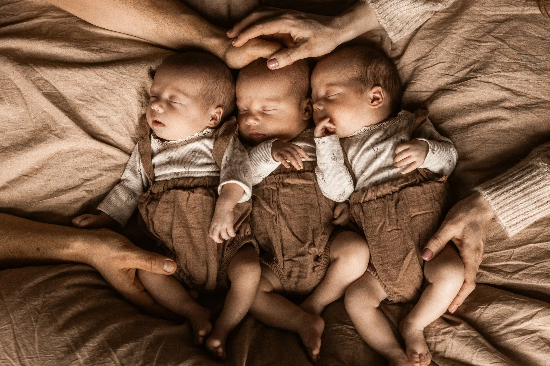 Tata z mamą otulają rękami trójkę noworodków śpiących na stylizowanym beanbag w kolorze beżowym podczas sesji zdjęciowej w domu