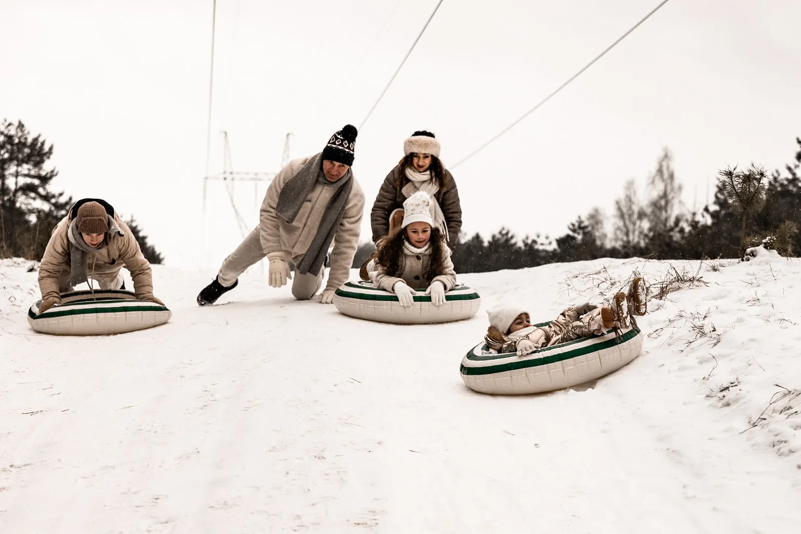 Dzieci zjeżdżające z górki podczas zimowej sesji zdjęciowej, rodzice w tle spoglądają na dzieci zjeżdżające z górki śnieżnej