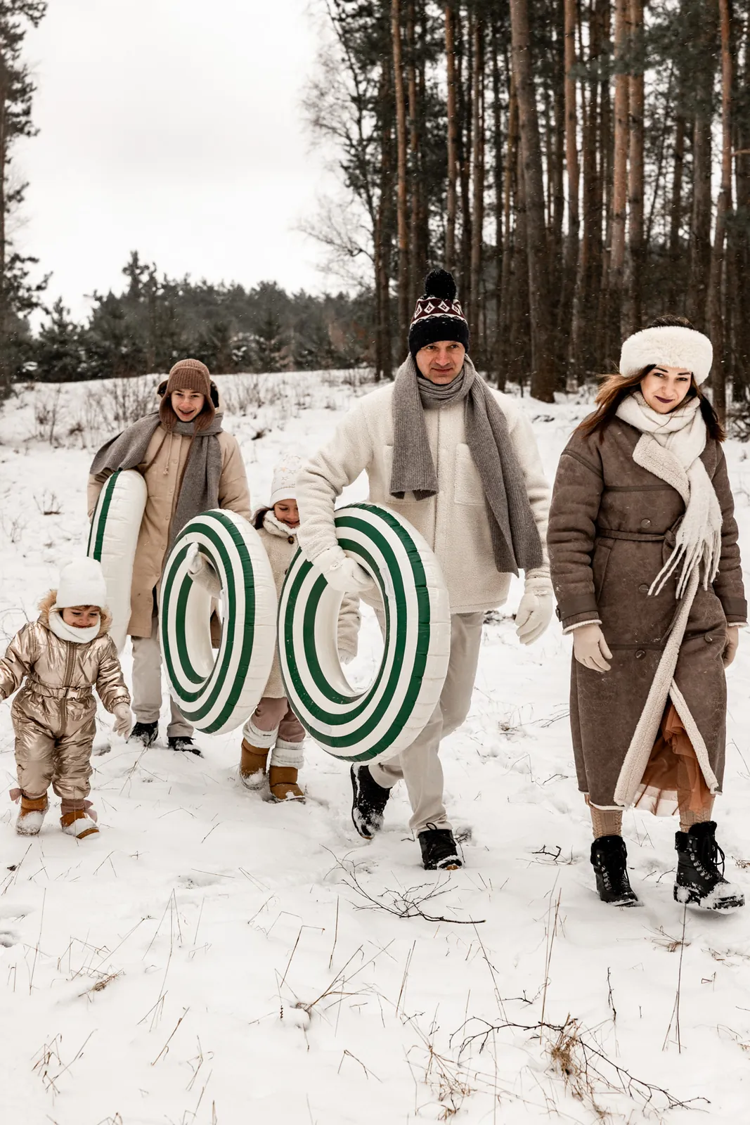 Rodzice z dziećmi podczas spaceru na śnieżna górę, trzymają w rękach koła zamiast sanek, w tle las i filmowa sceneria